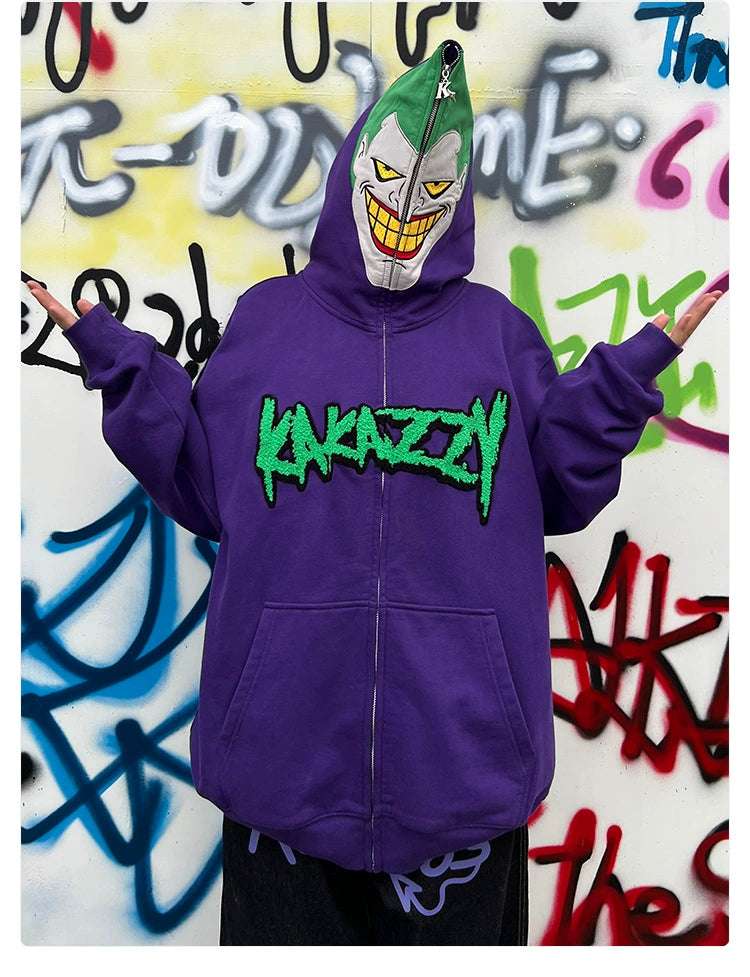 A person in a Seakoff Joker-Inspired Hoodie (400GSM Oversized Purple Zip-Up Supervillain Sweatshirt) stands with arms outstretched, hood up to hide their face behind a cartoonish mask. The backdrop features vibrant graffiti splashes.