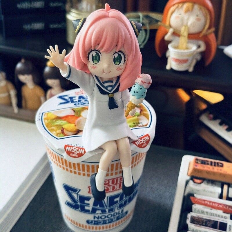 Anya Forger with Ice Cream, a PVC anime collectible from Spy × Family, features a pink-haired figure in a sailor-style outfit perched on a Nissin Cup Noodles container. In the background, more anime collectibles and various items adorn the shelf.