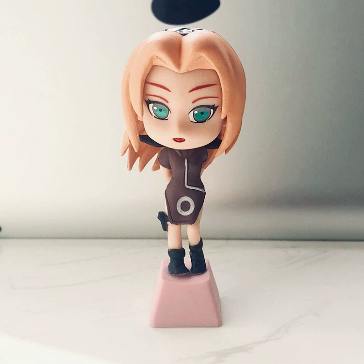 A small figurine of a character with long pink hair, green eyes, and a brown outfit stands on a pink base beside the sleek Ninja Masters Keycap Set from Naruto on a light surface. The figure has an expressive facial expression and is positioned in a slight tilt.