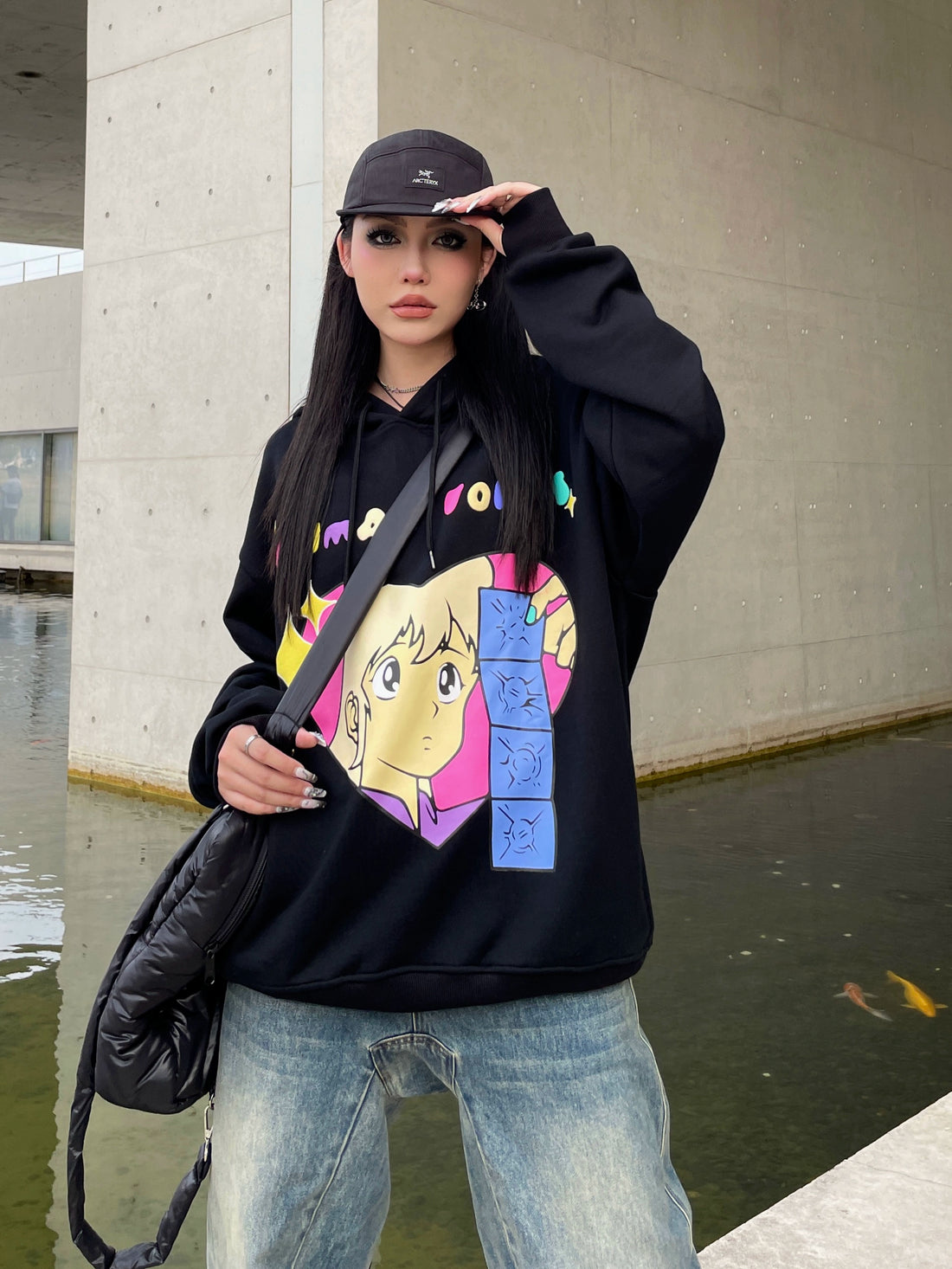 Wearing a Seakoff Retro Anime Hoodie – Human Voices Graphic Pullover, paired with blue jeans and a black cap, a person with long dark hair and a crossbody bag stands outdoors. With the backdrop of a concrete wall and water, the scene exudes nostalgic vibes.