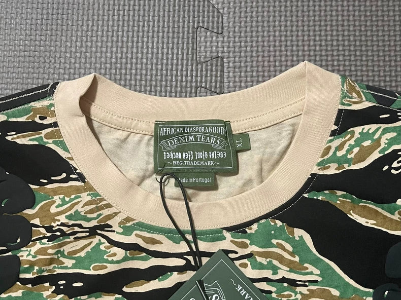 A close-up of the Denim Tears Green Camo All-Over Print T-Shirt with a beige collar, cotton tiger-stripe camouflage, and tags reading African Diaspora Goods, Denim Tears, and Made in Portugal, laid flat on textured gray.