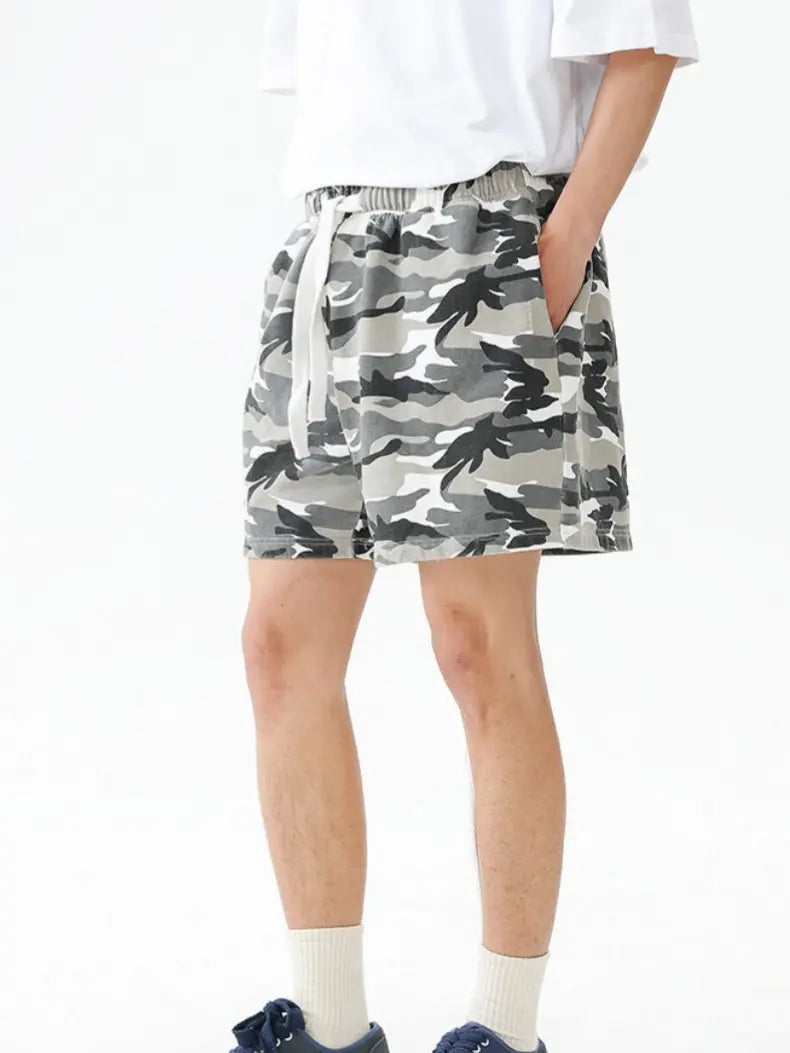 A person wears Men's Urban Camo Cotton Drawstring Shorts in gray camouflage, paired with white socks and dark sneakers. Only their lower body is visible, with hands tucked in pockets.