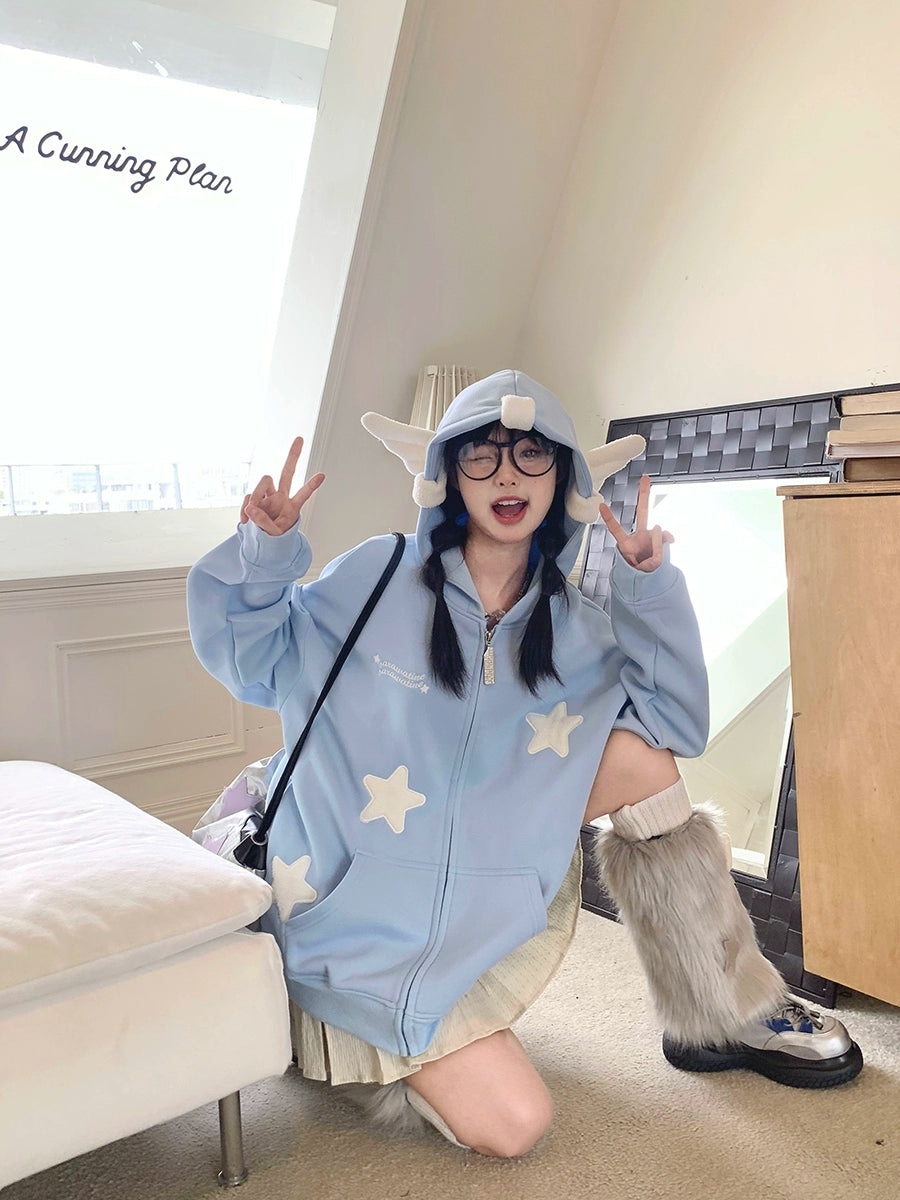 Indoors, a person showcases the Seakoff Kawaii Star Elf Hoodie – a cute 3D ear design with soft star appliques. They wear glasses and pose playfully on one knee with a peace sign. The softly lit room, featuring a white couch and wooden cabinet, enhances the kawaii-inspired fashion scene.