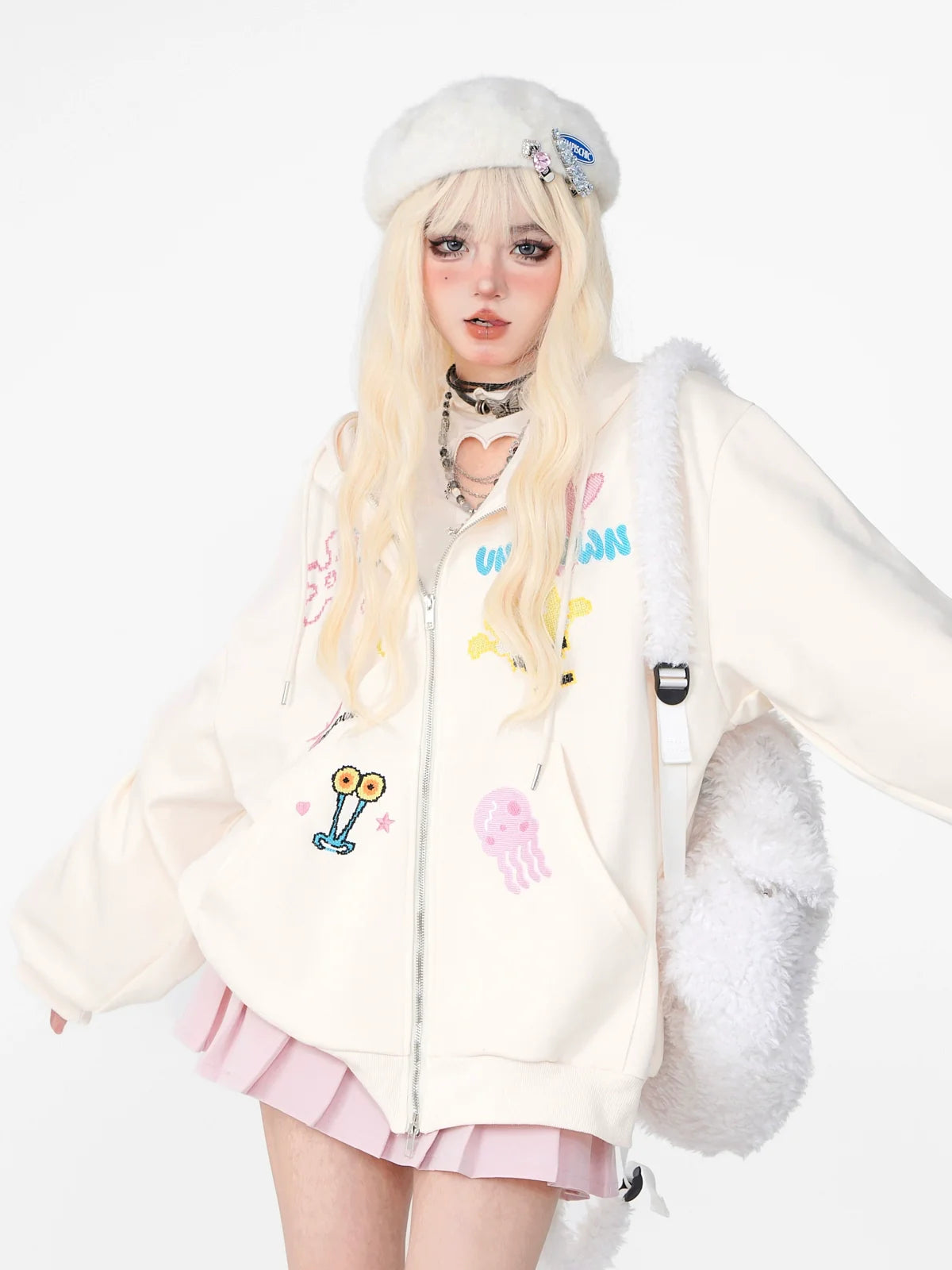 With long blonde hair, a person wears Seakoff's Kelly Kitty Pastel Kawaii Hoodie, accessorized with a white beret, faux fur shoulder bag, choker, and pink pleated skirt. The embroidered patches on the oversized hoodie highlight kawaii fashion against the plain white backdrop.