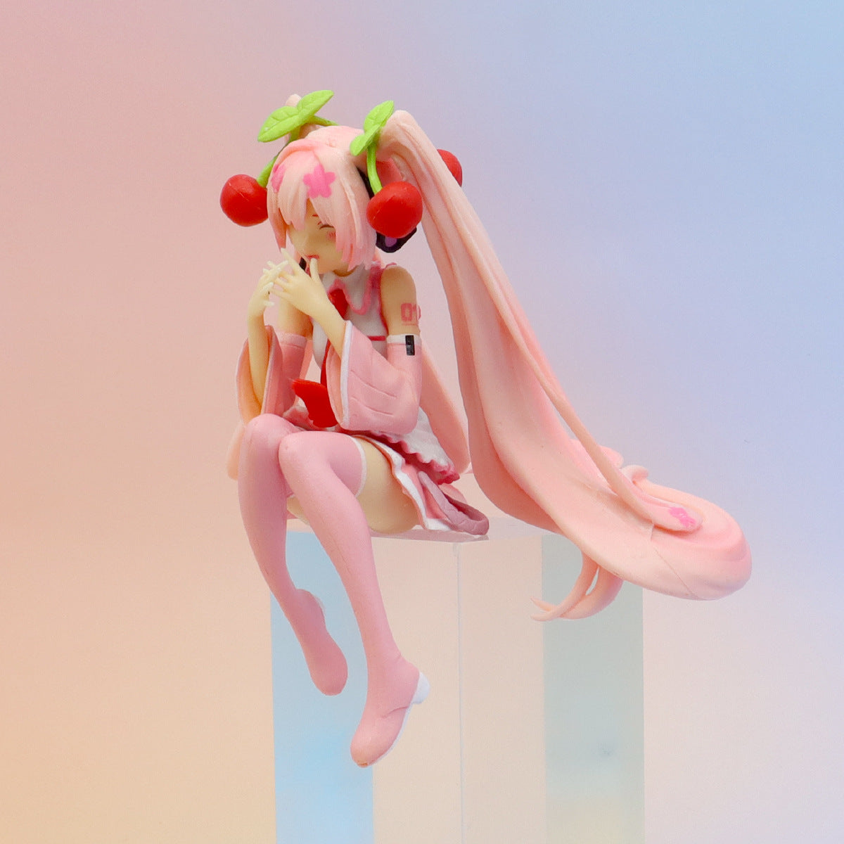 The Hatsune Miku Cherry Sitting Pose 11cm PVC Figure features an anime-style character with long pink hair and cherry embellishments. She sits gracefully with a smiling expression, hands near her face, wearing a pink and white outfit against a pastel gradient backdrop.