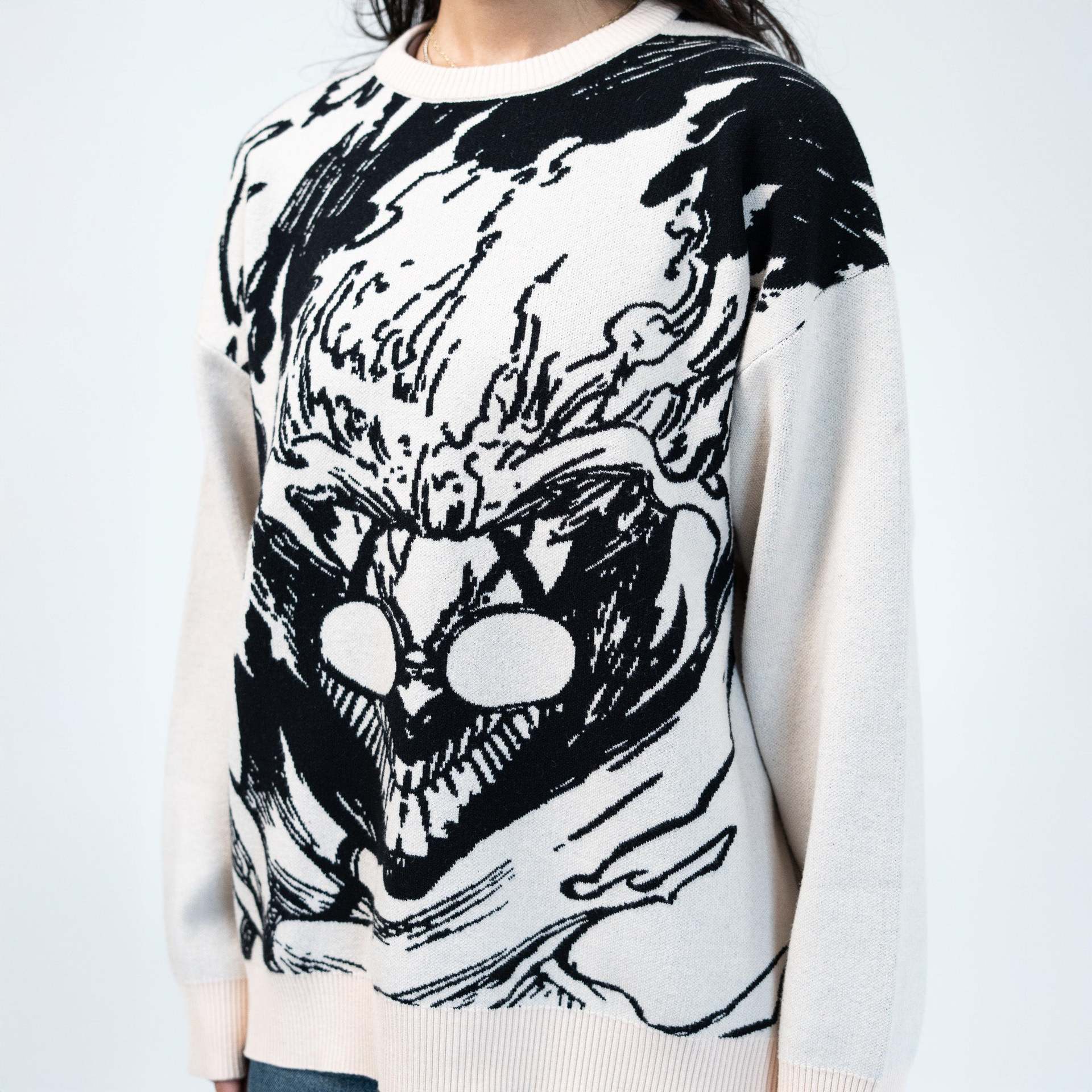 A person is wearing a Dandadan Premium Turbo-Granny Possessed Knit Sweater, featuring a large, abstract black and white graphic inspired by anime art. The jacquard design with bold lines and shapes creates a dynamic visual pattern on a beige background.