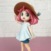 The Spy × Family Anya Forger Beach Day PVC Figure is a 17cm anime character with pink hair, a brown wide-brimmed hat, light blue dress, and dark blue shorts, standing against a light brick wall on a pink surface.