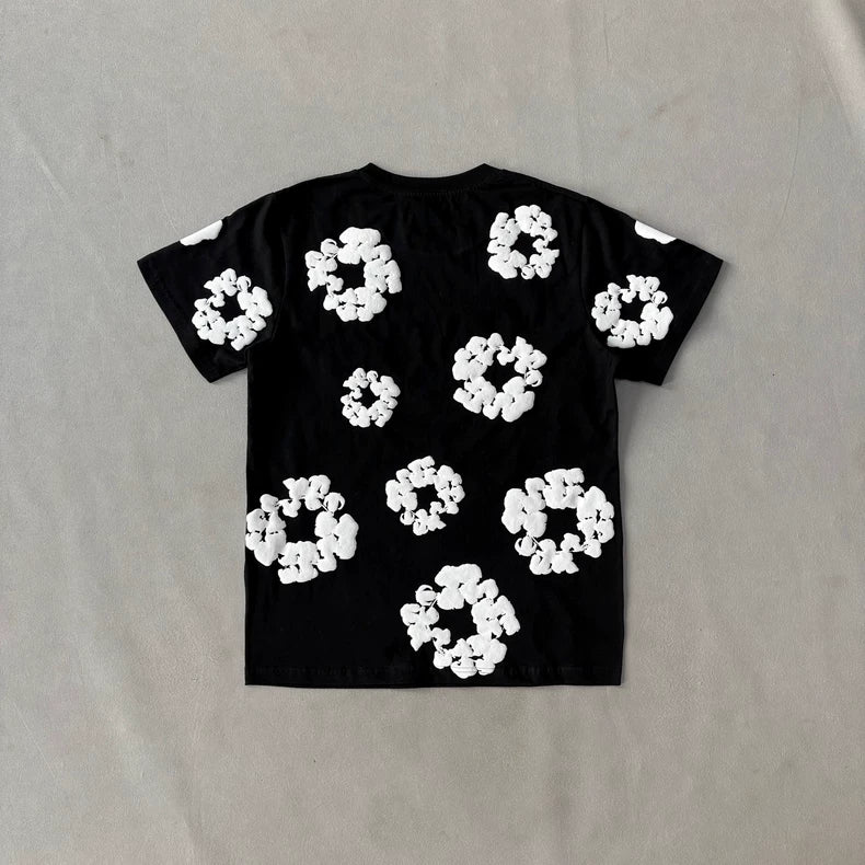 The Denim Tears Black Cotton Wreath All-Over Print T-Shirt is laid flat on a light surface, showcasing modern streetwear style with large and small white wreath-like patterns scattered across the back.