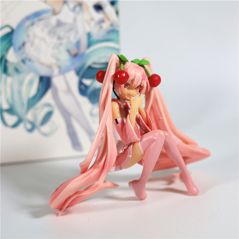 The Hatsune Miku Cherry Sitting Pose 11cm PVC Figure presents an anime character with long pink hair adorned with cherries, dressed in a pink outfit and knee-high boots, sitting gracefully on one knee with her hand near her mouth. A partially visible poster adds intrigue to the backdrop.