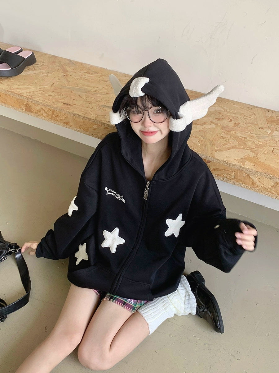 A person sits on the floor in the Seakoff Kawaii Star Elf Hoodie—featuring black fabric, white 3D ears, and soft star appliques—exuding a kawaii style. They have glasses and are smiling. Behind them is a wooden bench with shoes casually scattered on the floor.