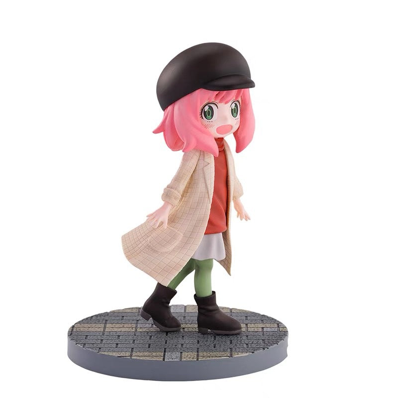 This 14 cm Spy x Family Anya Forger PVC figure features a cute chibi version with pink hair, wearing a black hat, beige coat, red dress, and green leggings. With wide eyes and an open mouth, it stands cheerfully on a textured round base.