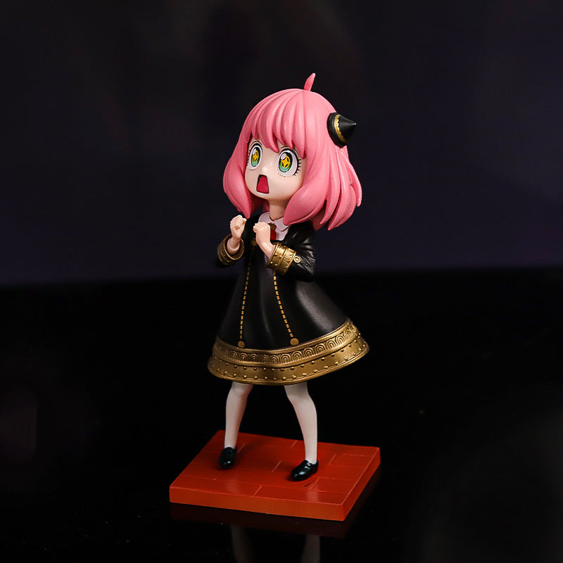 The Spy x Family Anya Forger PVC Figure features a chibi-style design with pink hair and a starry eyes expression. She wears a black dress with gold accents, stands on an orange base, and has small horn-like attachments on her head, set against a dramatic dark background.