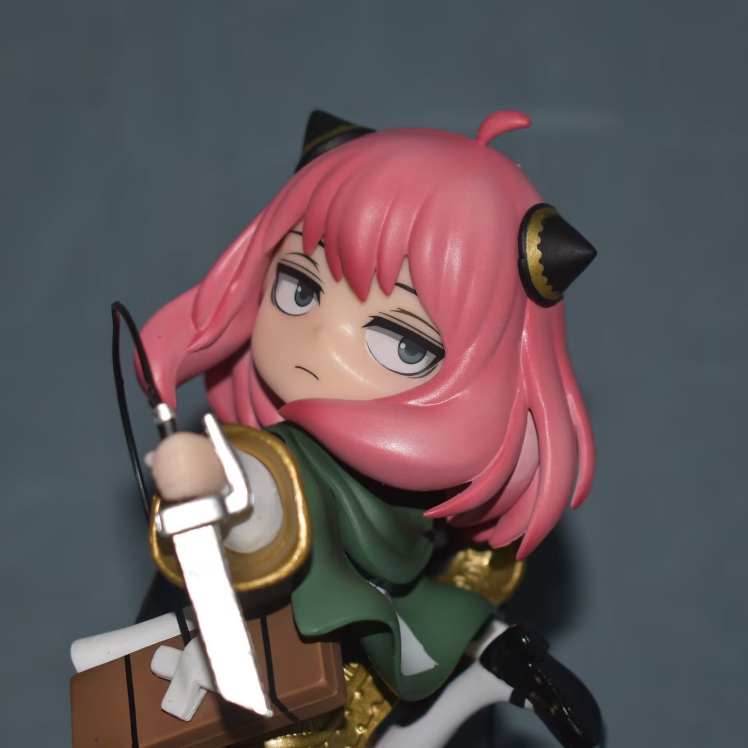 The Anya Forger Cosplay Figure by Spy × Family stands at 17.5CM, featuring a character with pink hair and horn-like accessories, posed with a sword. Clad in a green and gold outfit against a muted blue-green backdrop, this cute anime collectible is ideal for any merchandise collector.