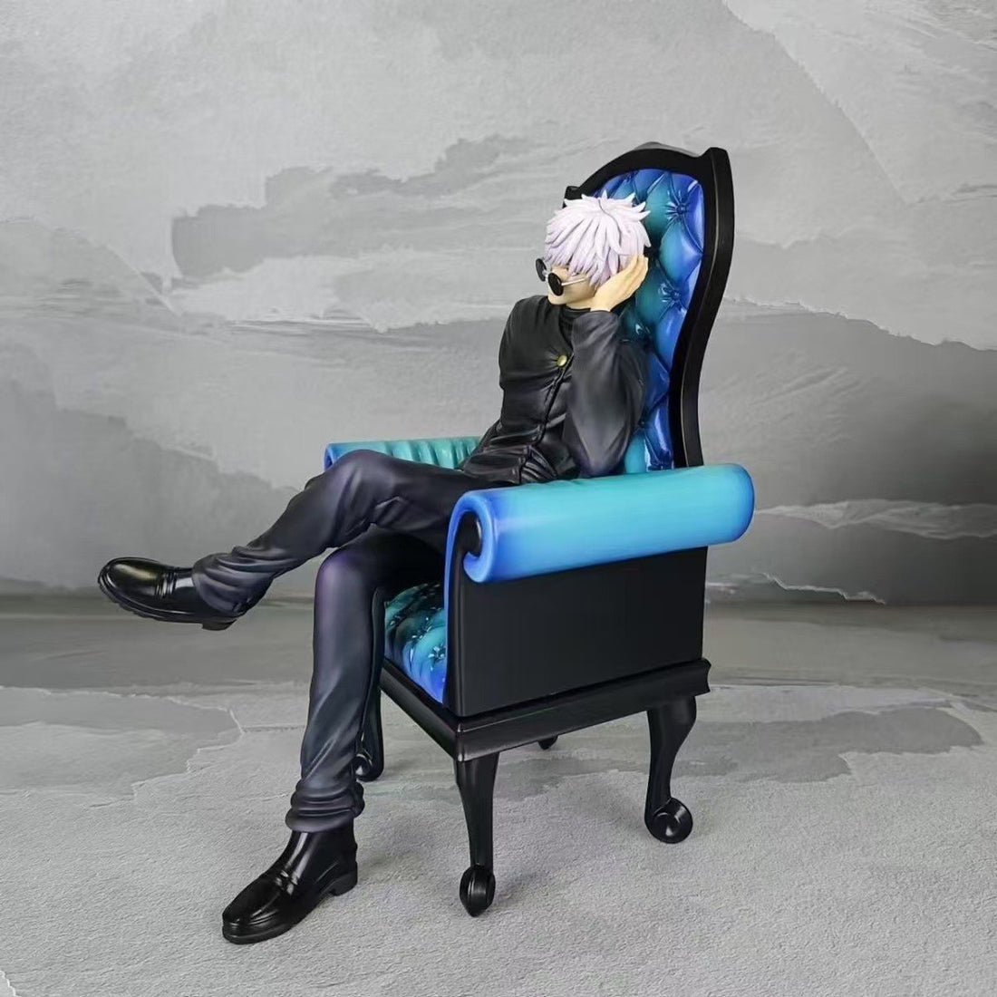 This Jujutsu Kaisen Satoru Gojo &amp; Suguru Geto Collectible PVC Figure Set features a white-haired figure in sunglasses, resembling Satoru Gojo, wearing a black outfit and seated stylishly in a black and blue armchair against a textured gray wall.