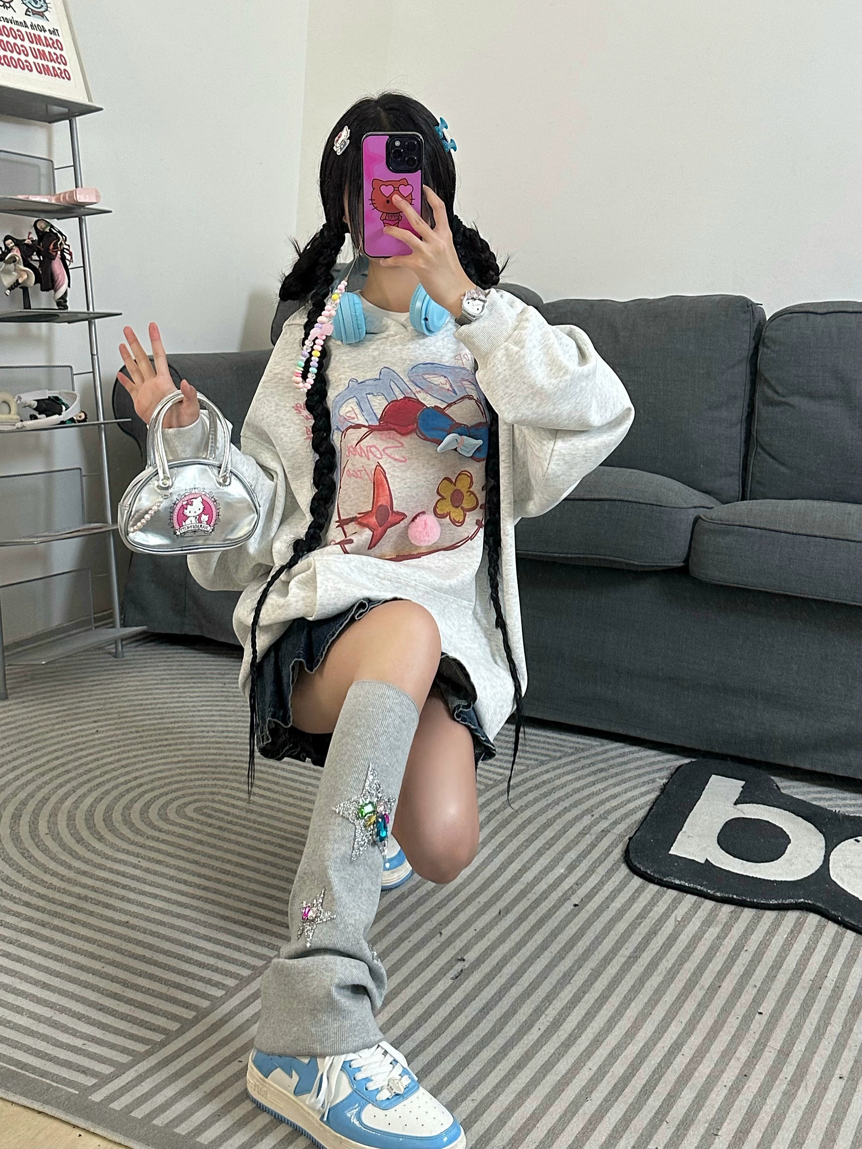 Kneeling on a patterned rug in front of a gray couch, they snap a selfie dressed in streetwear style—a Seakoff Kawaii Pom-Pom Crown Hoodie with unique cat graphics and 3D pom details. Gray leg warmers, sneakers, and a clear handbag enhance the kawaii look with colorful hair accessories.