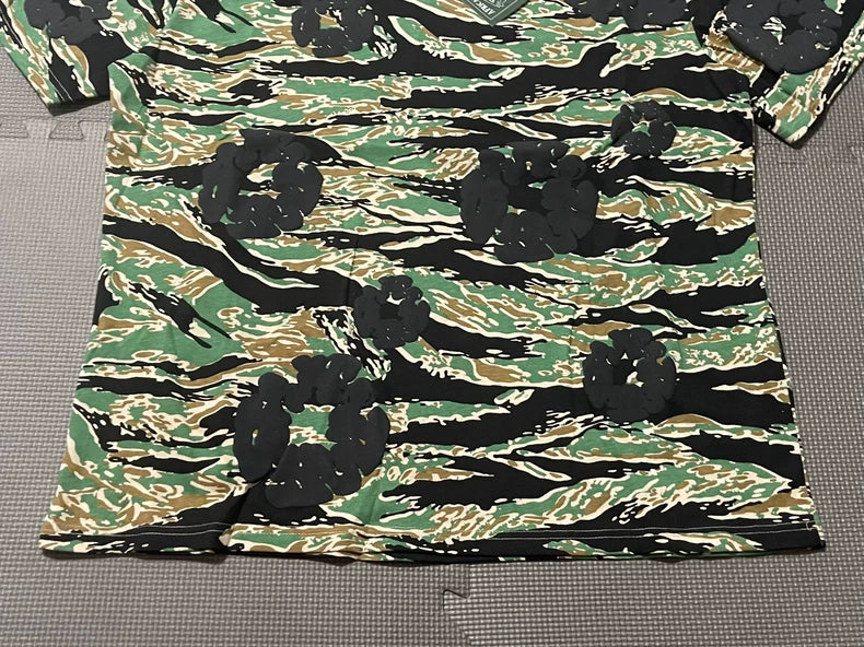 The Denim Tears Green Camo All-Over Print T-Shirt features green, brown, black, and tan tiger stripe camouflage patterns with bold black floral designs, styled flat on a textured gray background.