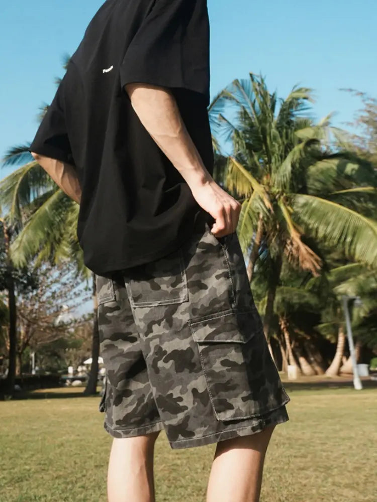 A person stands on grass with palm trees behind, wearing a black t-shirt and Men's Baggy Camo Cargo Shorts with Elastic Drawstring Waist and Flap Pockets in gray camo under a clear blue sky.