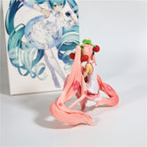 A charming Hatsune Miku Cherry Sitting Pose 11cm PVC Figure, with long pink hair and cherry accessories, gracefully poses in front of vibrant artwork. The figure features a pink outfit with delicate white details, capturing the iconic appeal of Hatsune Miku&