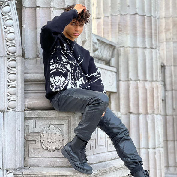 A person with curly hair, embracing streetwear style, sits on a stone ledge clad in a black Attack on Titan Premium Knit Sweater featuring an Eren Yeager Titan Form Jacquard Design, paired with black pants and boots. They gaze at the camera with one hand on their head amidst decorative stonework.