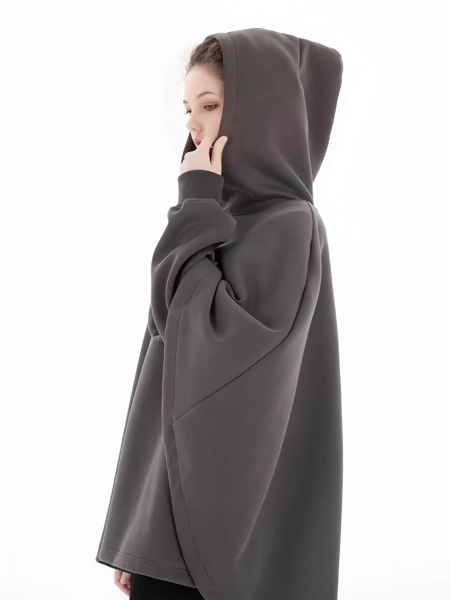 A person in a dark, oversized Limited Edition Attack on Titan Hooded Cape by Attack on Titan stands sideways against a white background, illuminated from the right with their hand near their face.