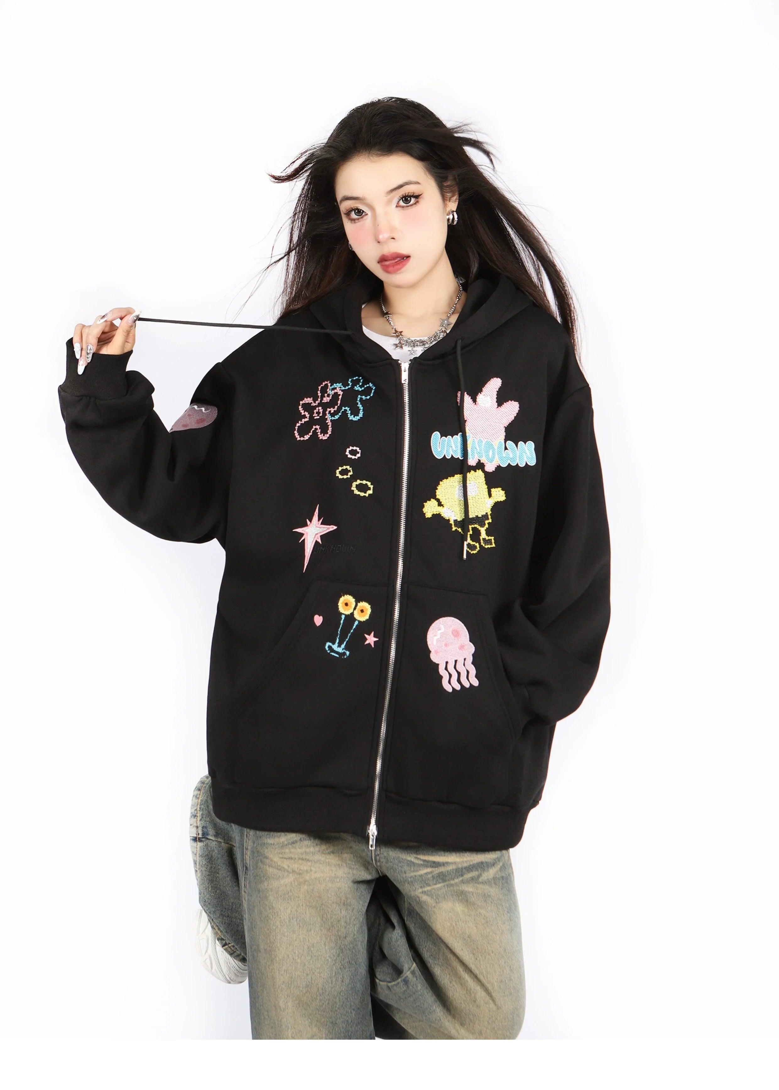 A person wears a Seakoff Kawaii Pastel Cartoon Hoodie, featuring playful embroidered designs like stars, planets, and flowers. They're paired with jeans against a white background, holding the hood's drawstring in an anime-inspired fashion.