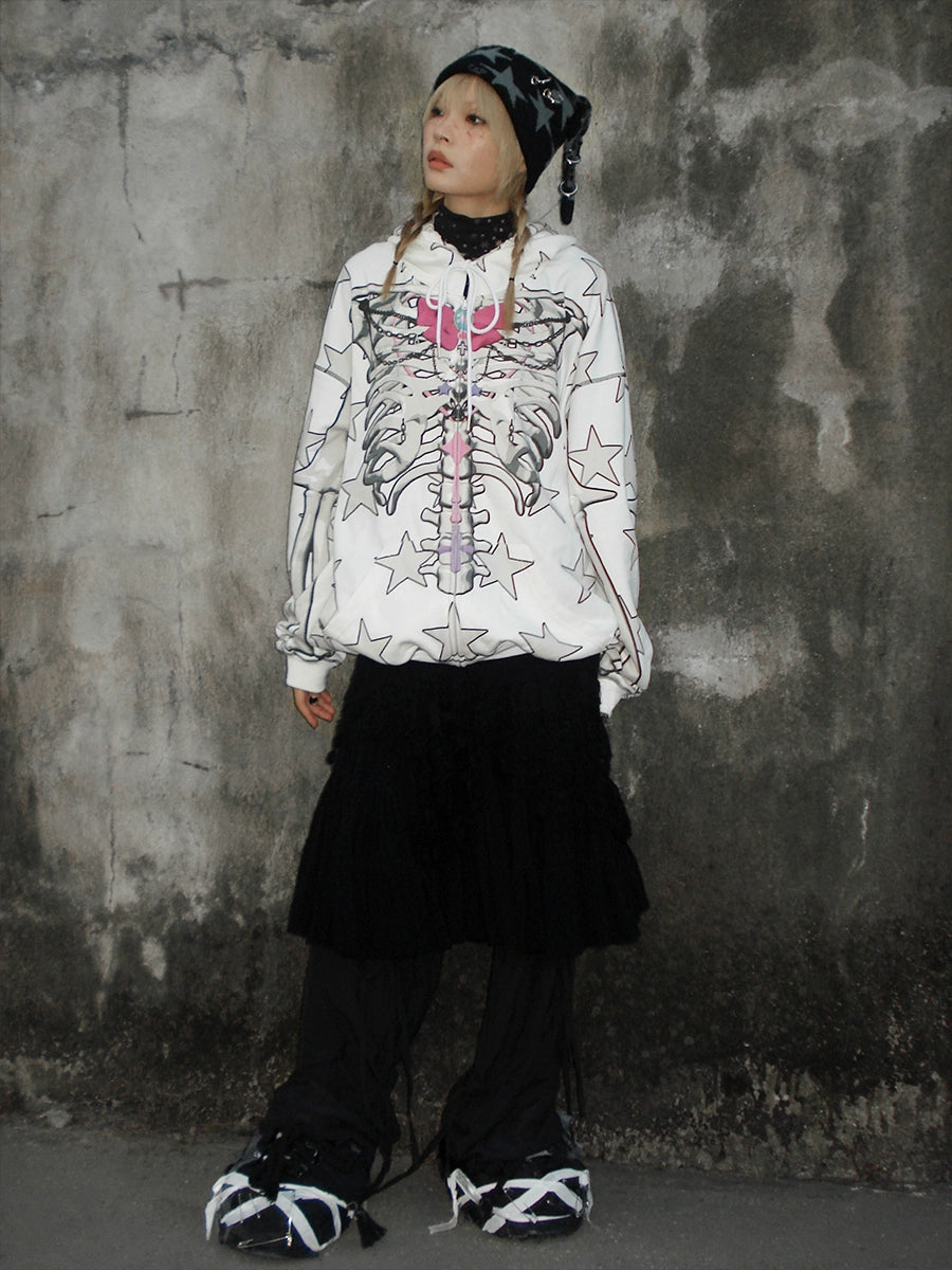 Against a concrete wall, a person sports the Seakoff Gothic Skeleton Star Hoodie—featuring a full-zip design with skeleton and star prints. A black beanie, skirt, and pants enhance their look, while shoes boasting unique white-black patterns complete their casually stylish pose.