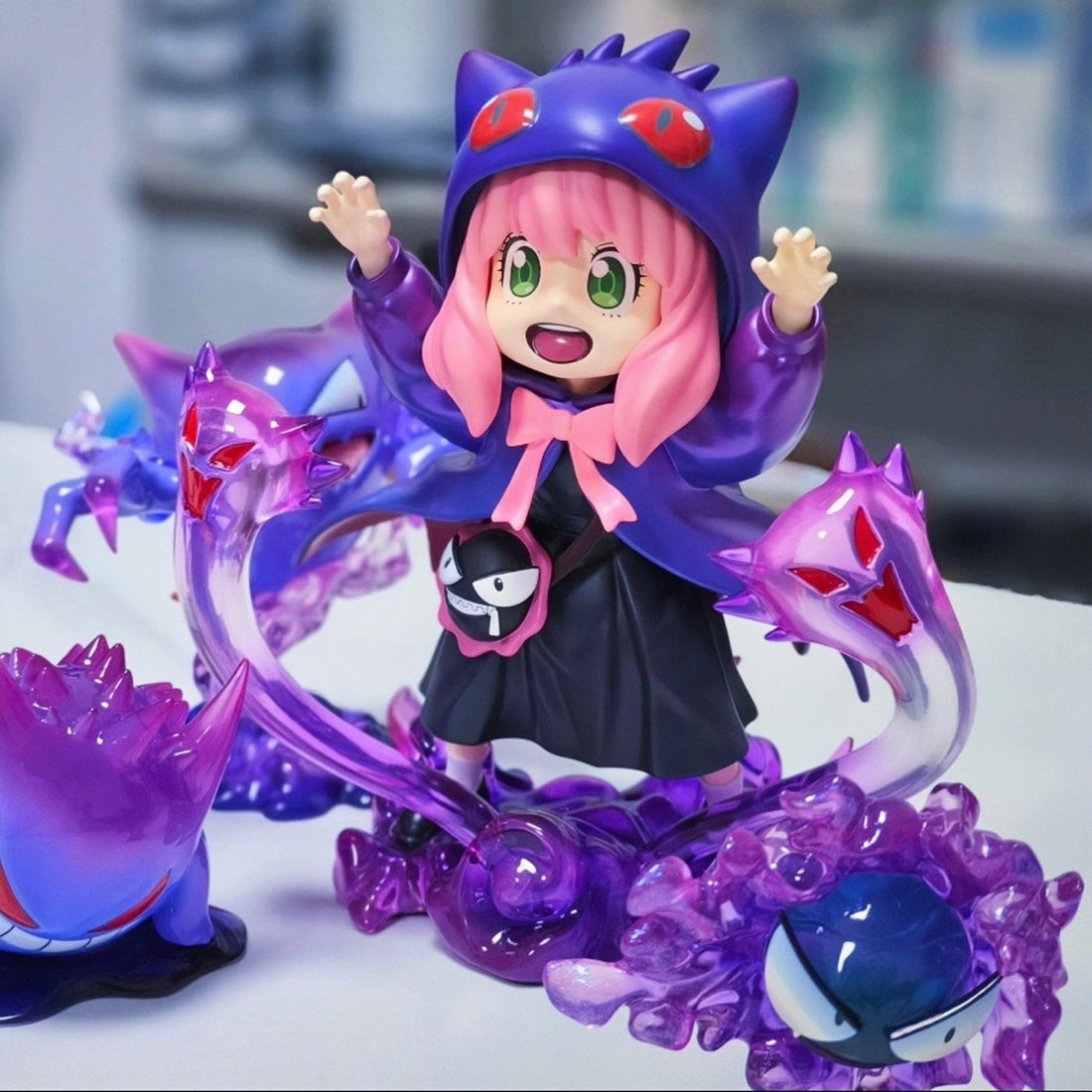 The Spy x Family Anya Forger PVC Figure, a 17 cm collectible, showcases Anya with pink hair and green eyes dressed in a purple Gengar cosplay. The figure features cat-like ears and red eyes surrounded by ghostly purple forms on a white base. Available in black &amp; white options.