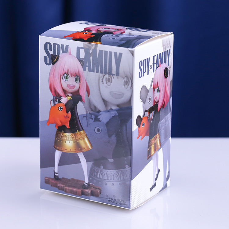 The Anya Forger Chainsaw Cosplay Figure features Anya with pink hair holding an orange creature. &quot;Spy × Family&quot; text and images of her in various poses decorate the box against a blue curtain and white tabletop background, making it an essential collectible for fans.