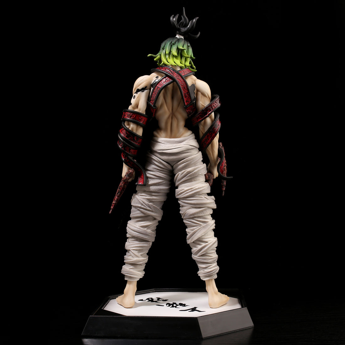 A Gyutaro Demon Slayer figure stands back-facing, showcasing white loose pants and red/black straps on the body. With green spiky hair, this Kimetsu no Yaiba collectible from the Demon Slayer brand rests on a base with Japanese characters.