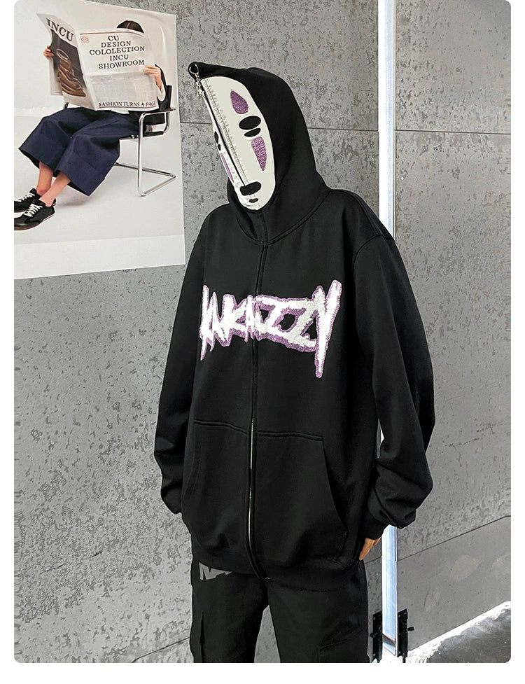 Wearing a black No-Face Anime Hoodie—a 400GSM oversized Spirited Away zip-up sweatshirt—a person stands against a metallic gray wall with a white mask. A poster with an anime character sitting is partially visible in the background, appealing to anime fans.