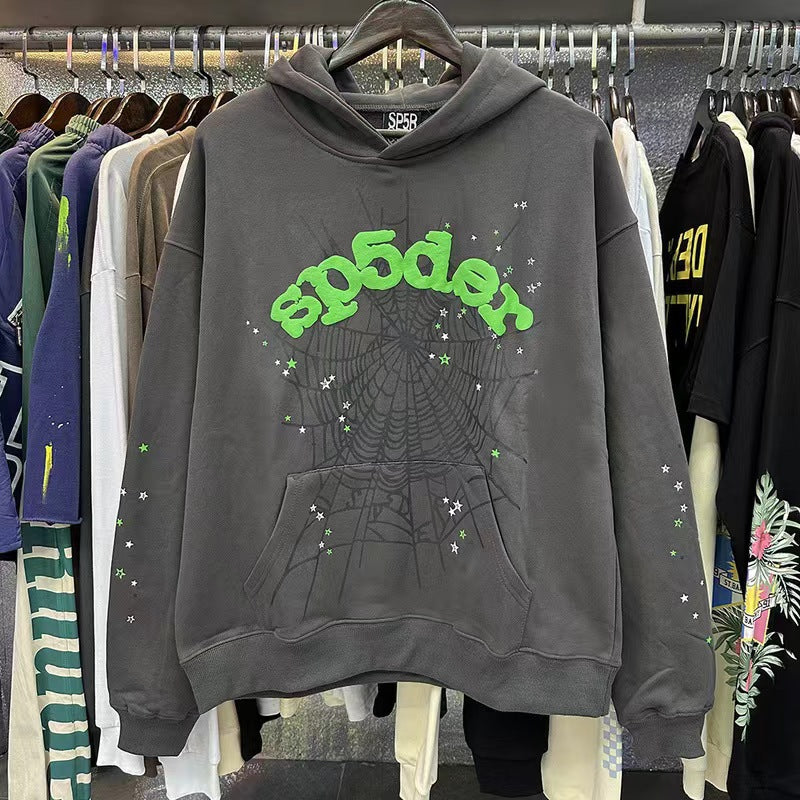 A Modern Grey Sp5der Hoodie by Sp5der hangs on a rack, featuring green graffiti-style &quot;sp5der&quot; text, a web print design, and small stars, standing out among other streetwear items in a bustling retail setting.