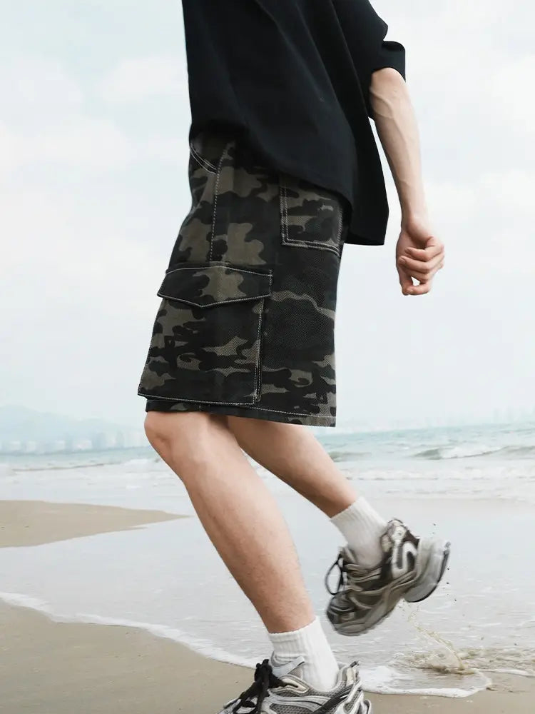 A man in a black shirt, white socks, gray sneakers, and Men's Baggy Camo Cargo Shorts with Elastic Drawstring Waist and Flap Pockets walks along a sandy beach with gentle waves in the background.