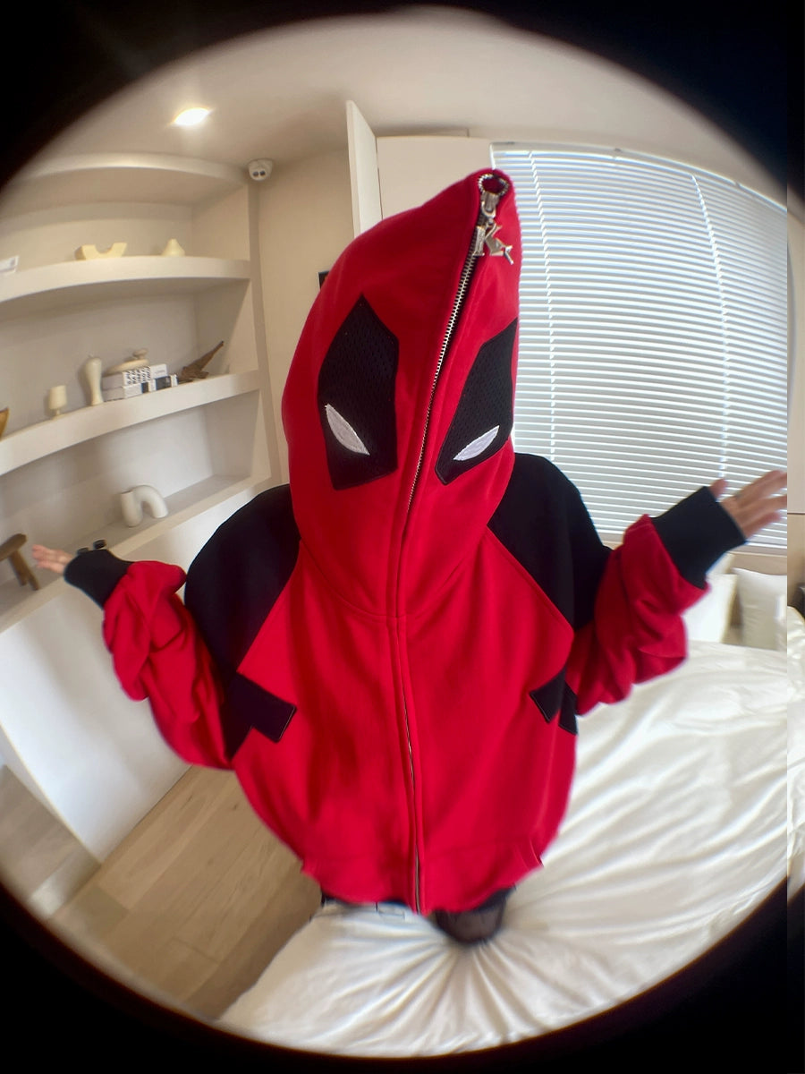 The person wears a Seakoff Deadpool-Inspired Hoodie, a 400GSM oversized red and black zip-up sweatshirt with a superhero mask design. They zip it up to obscure their face, standing indoors against shelves and window blinds, embracing a streetwear vibe.