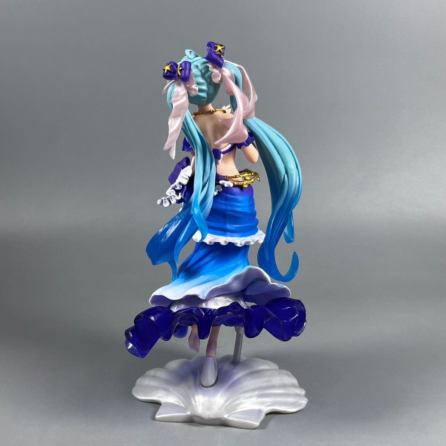 The Hatsune Miku Mermaid 24cm PVC Figure features the beloved character with flowing blue and pink hair, elegantly dressed in a blue and white layered dress. It poses on a seashell-shaped base against a gray backdrop, making it an elegant and stunning collectible for any Vocaloid fan!.