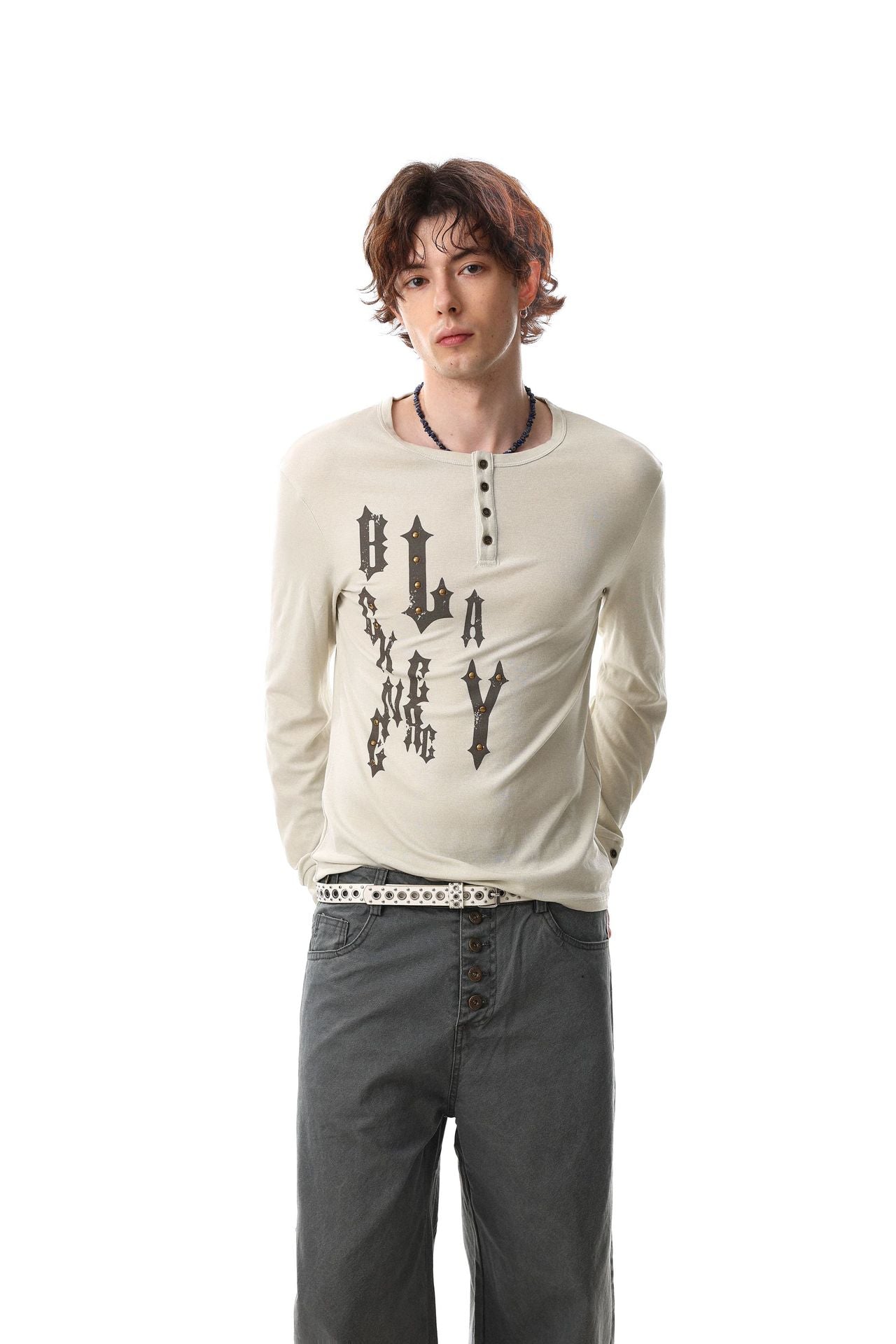 Chemise henley beige à manches longues, motif clouté en lettrage blackletter gothique sur la poitrine, coupe ajustée, portée par un mannequin avec un jean à jambes larges