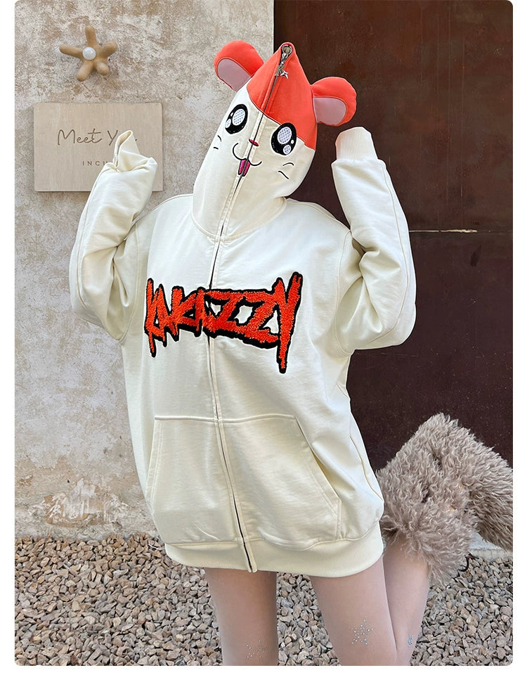 A person models the Seakoff Cute Hamster Cartoon Hoodie, crafted from 400GSM cotton. This charming hoodie features a playful ear design, a face on the hood, red graphic text on the front, and a fluffy tail accessory. Ideal for cartoon lovers, they are pictured standing outdoors on gravel.