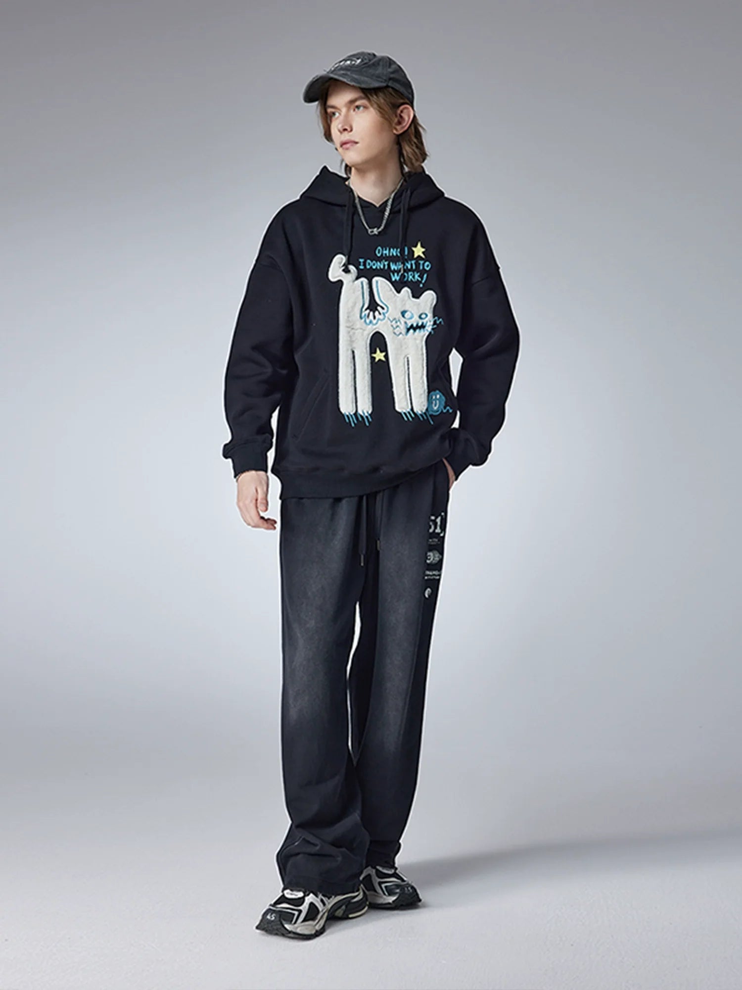 A person stands against a gray background wearing Seakoff's "Oh No! I Don’t Want to Work!" oversized hoodie featuring a lazy cat design, paired with dark pants, sneakers, and a baseball cap, perfectly embodying casual streetwear vibes.