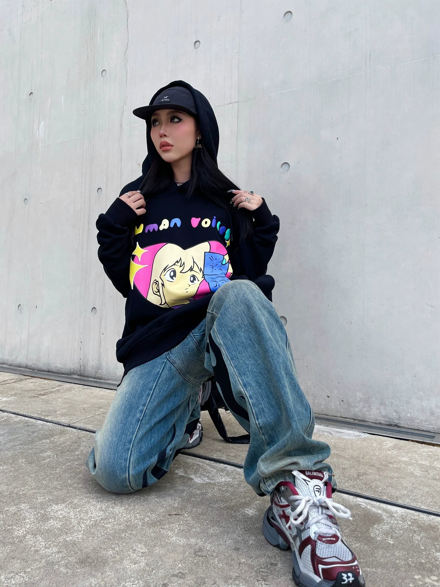 Kneeling on the ground, a person sports Seakoff's Retro Anime Hoodie—a Human Voices Graphic Pullover with a drop shoulder design—over blue jeans and sneakers. The hood is pulled over a black cap, evoking nostalgic vibes against a stark concrete wall.