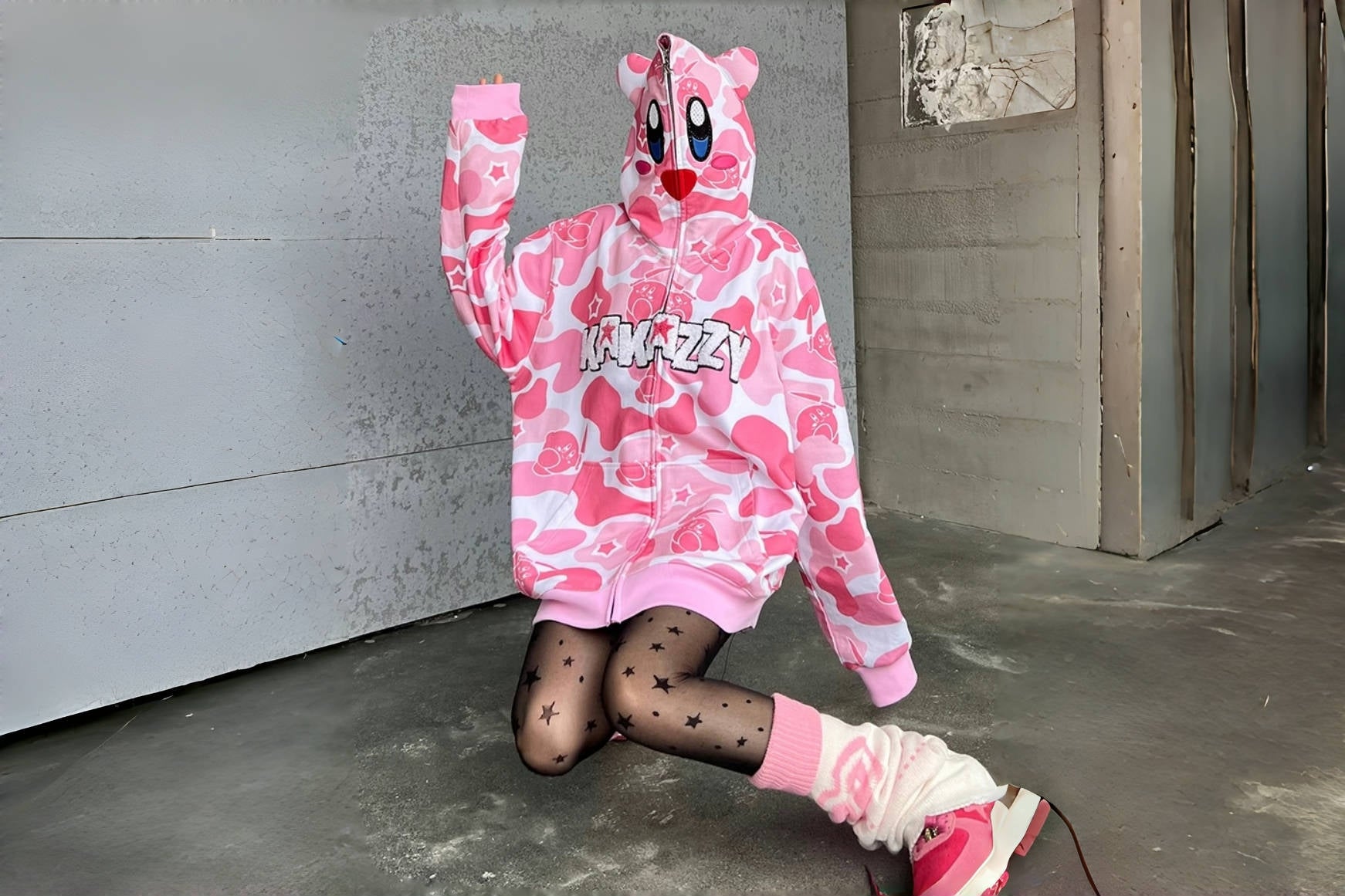Person kneeling on the ground wearing a pink hoodie with an animated face and the word "AMAZZY," patterned tights with stars, and leg warmers. The setting appears to be an industrial or urban space with concrete walls.