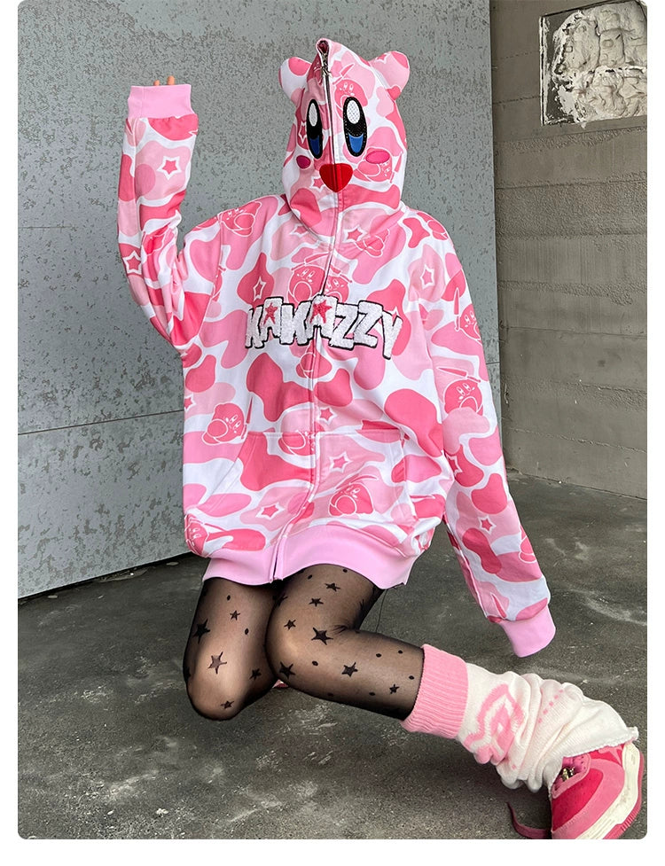 A person wearing a Seakoff Pink Kirby-Inspired Hoodie, featuring a camo design and 3D ears, sits with the hood up. They&