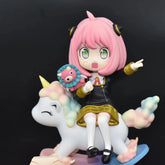 The Spy x Family Anya Forger PVC Figure (15 cm) features a pink-haired girl with interchangeable expressions riding a colorful unicorn with a rainbow mane and tail. She holds a plush toy and points, set against a plain black backdrop.