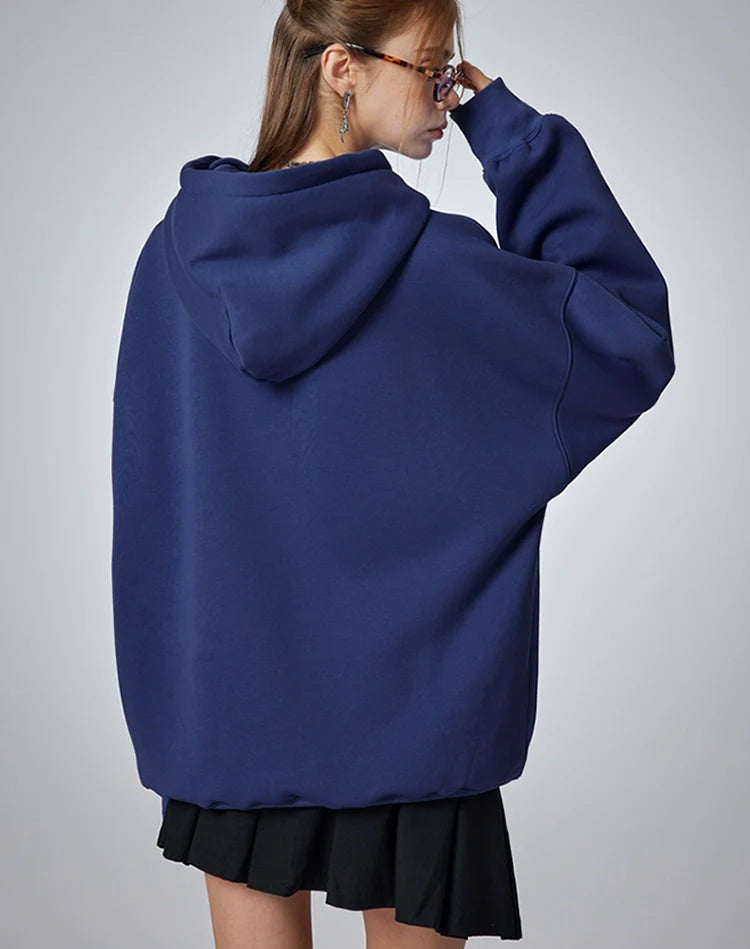 A person with long hair, seen from behind, wears a Seakoff "Oh No! I Don’t Want to Work!" oversized hoodie with a lazy cat design and a black skirt. They adjust their glasses with one hand near their face against a neutral gray background, enhancing the casual streetwear vibe.