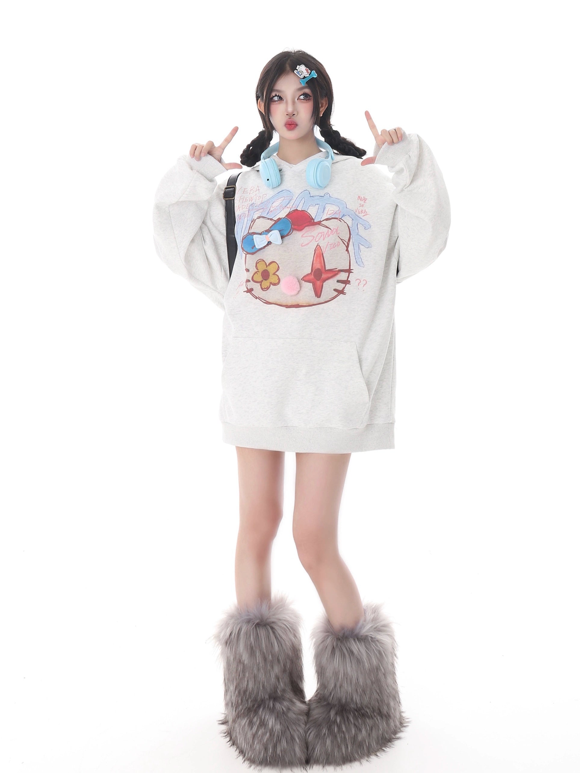 Against a white background, a person embraces streetwear in the Kawaii Pom-Pom Crown Hoodie by Seakoff, featuring a unique cat graphic. They wear fluffy gray leg warmers and have headphones around their neck. Colorful accessories adorn their braided hair as they gesture upward with both hands.