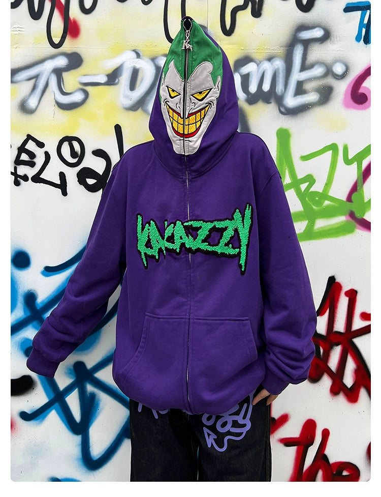 Someone in a Seakoff Joker-Inspired Hoodie, a 400GSM oversized purple zip-up featuring the word &quot;Krazy&quot; in green and a menacing face on the hood, stands against graffiti-covered walls, embodying supervillain style.