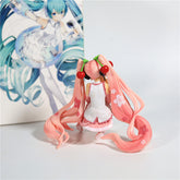 The Hatsune Miku Cherry Sitting Pose 11cm PVC Figure from the Hatsune Miku brand features long pink hair in pigtails with cherry accessories, flower motifs on her outfit, and includes an illustrated poster background.