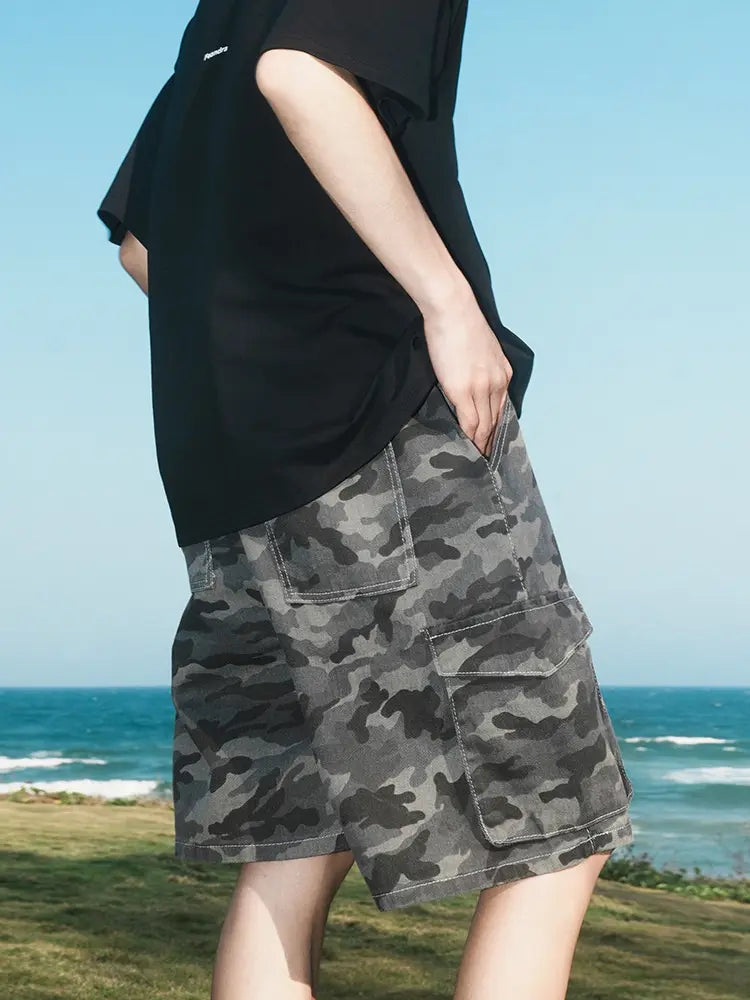 A person stands outdoors by the ocean, with green grass and blue sky behind, wearing Men's Dark Grey Camo Cargo Shorts with Elastic Waist and Flap Pockets and a black T-shirt.