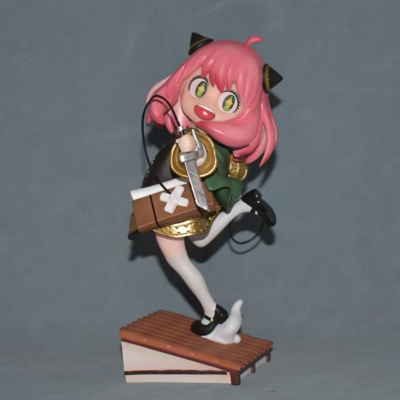 The Anya Forger Cosplay Figure from Spy × Family features a pink-haired character in a green outfit and black shoes, balancing on one leg with cat-like eyes, holding a brown bag against a gray backdrop—a charming 17.5cm anime collectible with a sword.