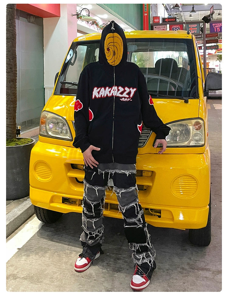 In front of a yellow vehicle, a person wears a Seakoff Uchiha Obito-Inspired Hoodie featuring an Akatsuki Cloud and spiral mask. Paired with black jeans with frayed patches and red and white sneakers, it&