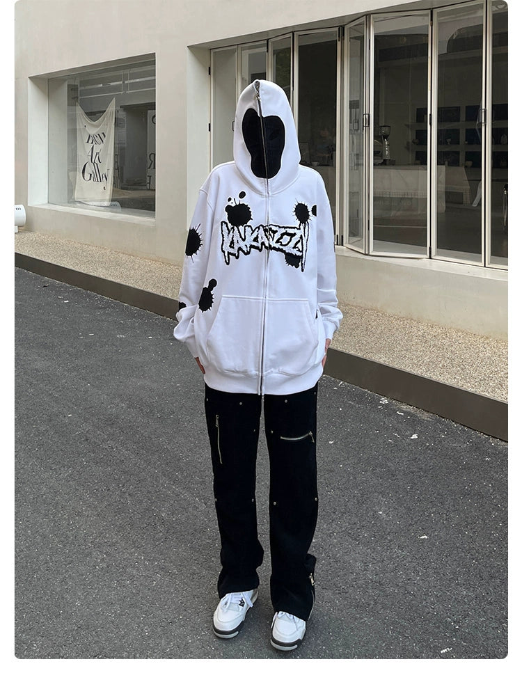 A person in front of a white building wears Seakoff’s Monochrome Ink Splatter Hoodie, a 400GSM oversized white and black zip-up sweatshirt, paired with black pants and white sneakers, creating an ideal streetwear look. The hoodie is up, obscuring the face.