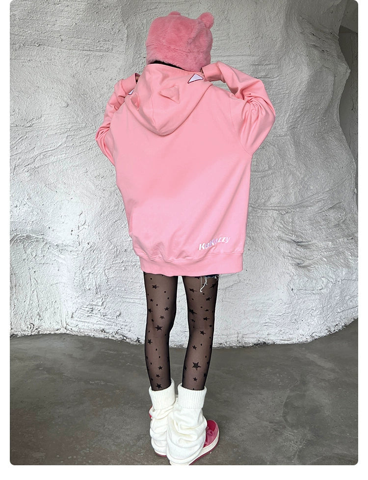 A person wearing a Seakoff Batman-Inspired Hoodie—an oversized pink zip-up superhero sweatshirt, black star-patterned tights, and white leg warmers—stands with their back to the camera. A pink pom-pom hat completes this fan&